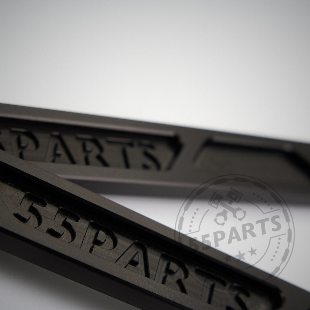 Nahaufnahme von zwei schwarzen 55Parts Exclusive Billet Aluminium Domstreben Vorderachsen passend für BMW 2er G42, 3er G2x und 4er G2x mit „55Parts“-Gravur; im Hintergrund ist ein verschwommenes BMW 55Parts-Logo sichtbar.