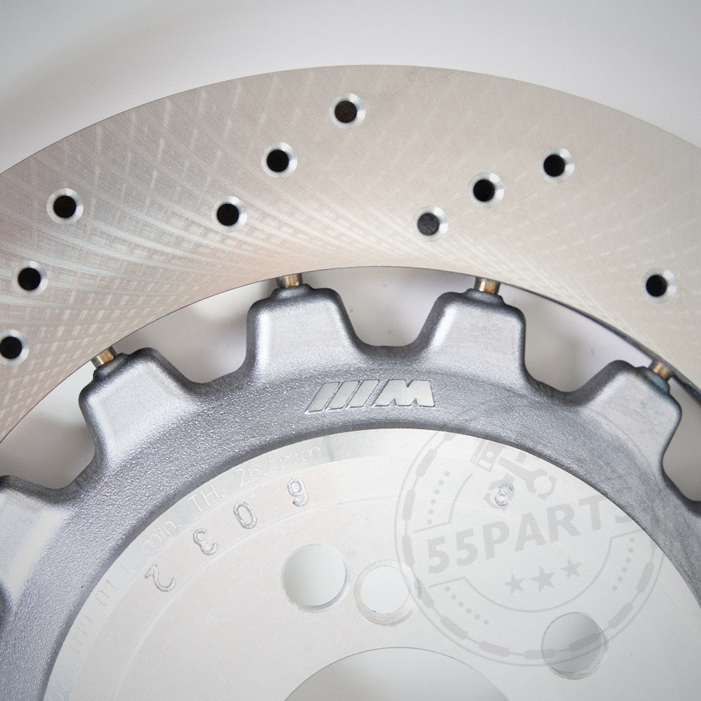 Nahaufnahme eines belüfteten und gebohrten 55Parts AP Racing CP9449 Big Brake Kit mit originaler 380x28 BMW-Scheibe passend für BMW M2, M3, M4 (F80, F82, F83, F87); im Hintergrund sind ein Teil eines Zahnrads und ein schwaches Logo sichtbar.