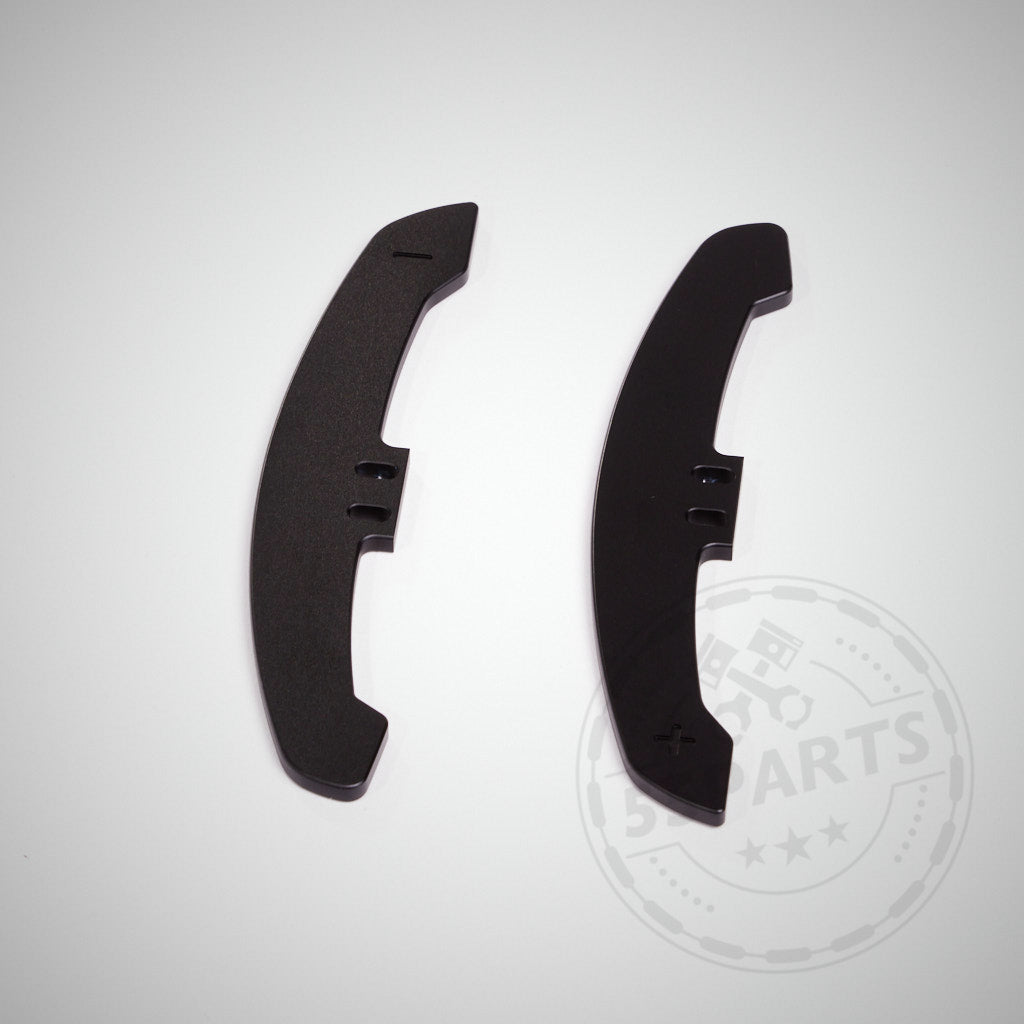 Zwei schwarze, gebogene CNC-gefräste Aluminium-Schaltwippen mit Plus- und Minus-Symbolen auf weißem Hintergrund, entwickelt für magnetische Schaltwippen für verbesserte Präzision.