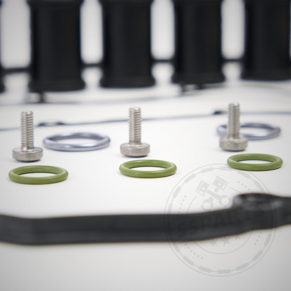 Nahaufnahme von grünen O-Ringen, Schrauben und Metallscheiben – Komponenten aus dem 55Parts Exclusive Einbauset Ventildeckel passend für BMW E9x M3 (S65 Motor) – auf einer weißen Oberfläche mit unscharfen schwarzen Teilen im Hintergrund.