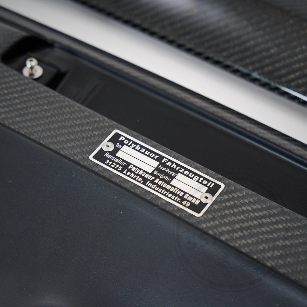 Nahaufnahme eines Metallschilds auf dem 55Parts Carbon Heckspoiler Heckflügel Verstellbar mit Teilegutachten für Toyota GR Yaris (inkl. Facelift), das Herstellerinformationen in deutscher Sprache zeigt.