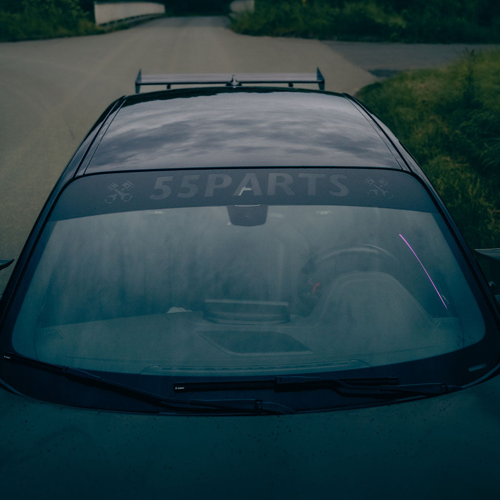 Ein dunkles Auto mit einem 55Parts-Scheibenkeil passend für alle Autos von 55Parts auf der Windschutzscheibe, in der Nähe von Gras geparkt und von oben gesehen, verwendet Oracal Markenfolie für eine blasenfreie Montage.