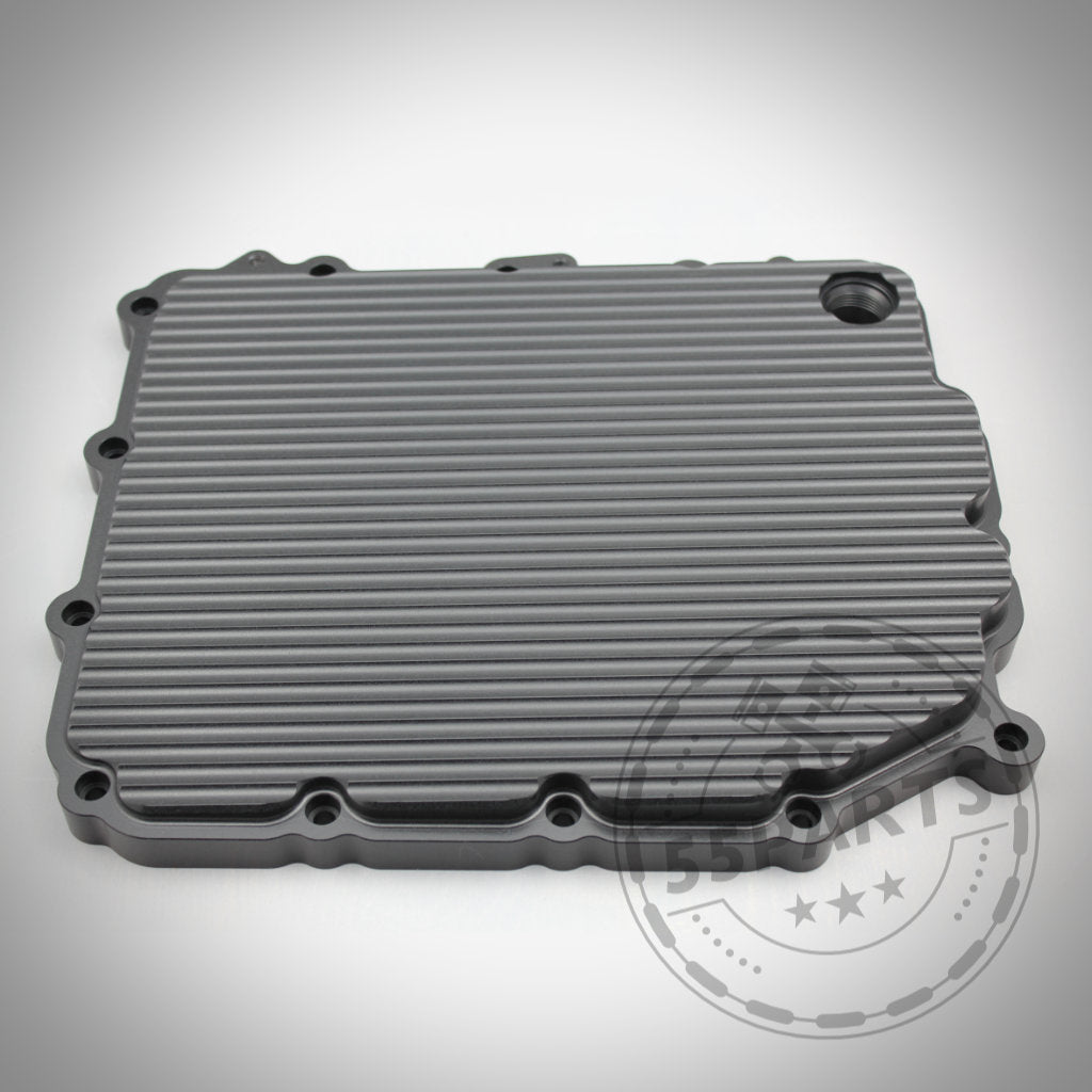 Die 55Parts Exclusive DKG GS7D36BG Aluminium-Ölwanne für die BMW 1/335i, M2(c), M3, M4, M5 und M6 E- & F-Serie verfügt über ein geripptes rechteckiges Design mit Schraubenlöchern und einer runden Öffnung, dargestellt auf weißem Hintergrund.