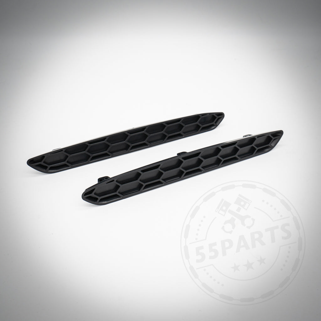 Zwei schwarze Reflektoren-Einsätze-Ersatz-Honeycomb-Teile, die für den BMW F87 M2 Competition entwickelt wurden, ruhen auf einer weißen Oberfläche mit einem dezenten 55Parts-Logo im Hintergrund.
