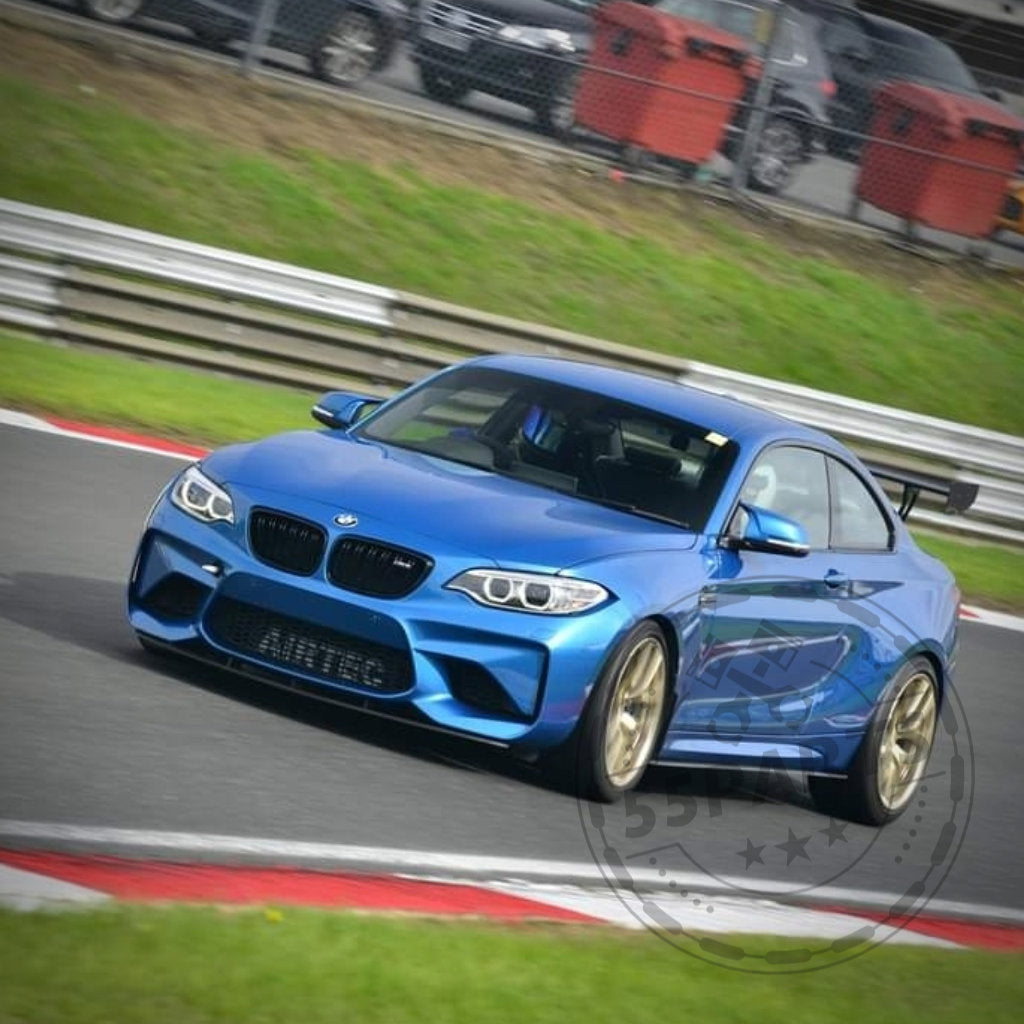 Ein BMW F87 M2 mit 55Parts Seitenschwellern aus Kunststoff PE Mattschwarz nimmt eine Kurve auf einer Rennstrecke, umgeben von Gras und Barrieren im Hintergrund.