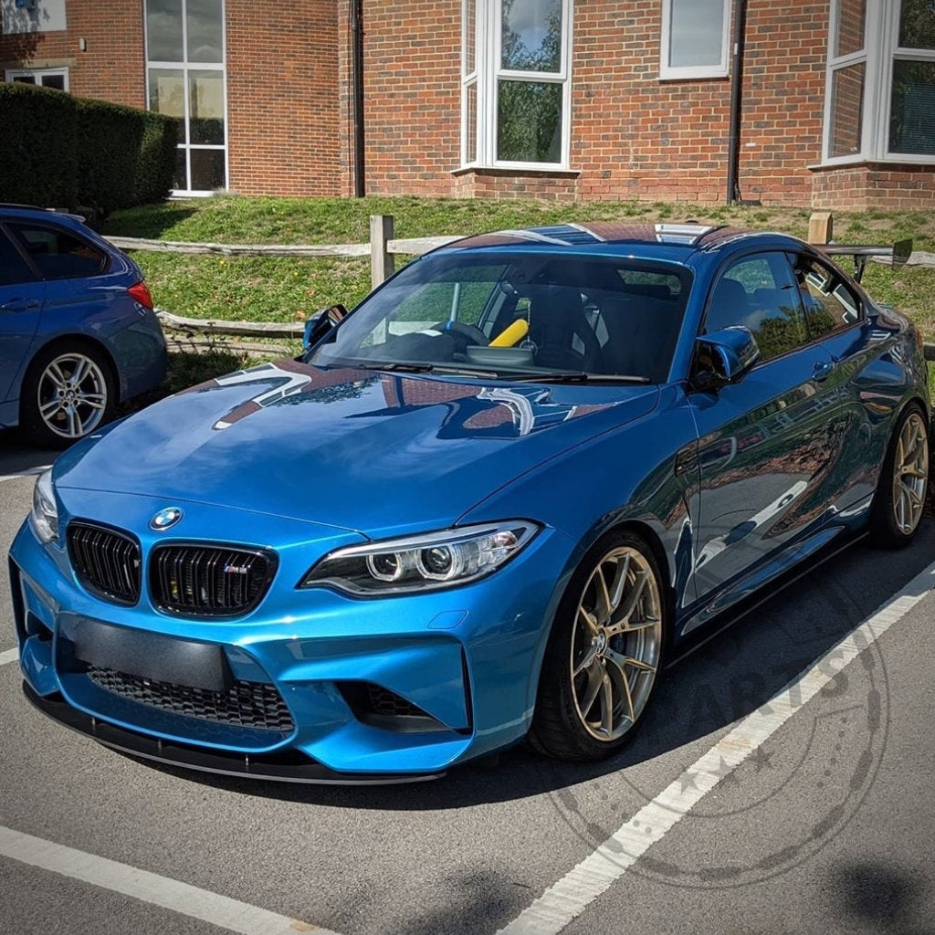 Ein blauer BMW F87 M2 mit 55Parts Kunststoff PE mattschwarzen Seitenschwellern steht an einem sonnigen Tag auf der Straße vor einem Backsteingebäude.