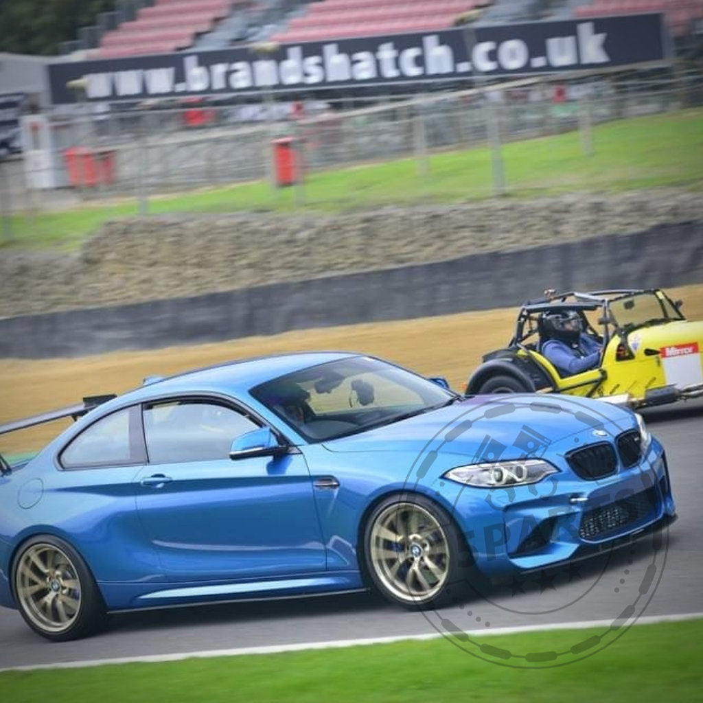 Ein blauer BMW F87 M2 mit 55Parts Seitenschwellern aus Kunststoff PE Mattschwarz und ein gelbes Open-Wheel-Autorennen auf der Rennstrecke von Brands Hatch, Hintergrund unscharf.