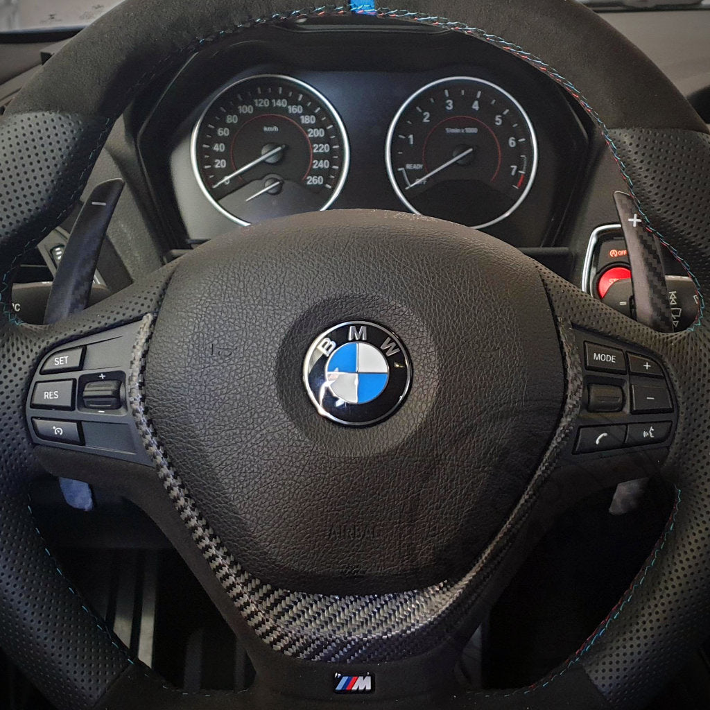 Nahaufnahme der 55Parts Aluminium/Carbon-Schaltwippen passend für BMW 1/235 & 40i, 3/435 & 40i, M2, M3 & M4 F-/G-Serie am Lenkrad mit sichtbaren Armaturenbrettanzeigen und Steuertasten.