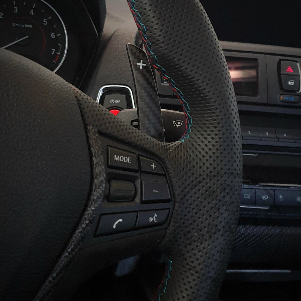 Nahaufnahme eines BMW-Lenkrads mit 55Parts Aluminium/Carbon-Schaltwippen für 1/235 und 40i, 3/435 und 40i, M2, M3 und M4 F- und G-Serie, mit Tasten und einem Armaturenbrett im Hintergrund.