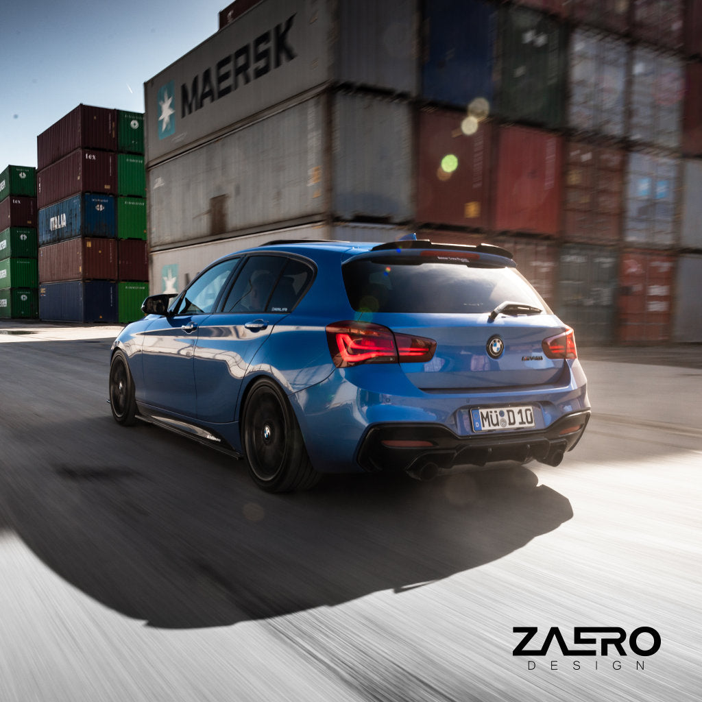 Ein blauer BMW M135i, ausgestattet mit einem ZAERO Evo-1 Diffusor, fährt im hellen Sonnenlicht an gestapelten Schiffscontainern vorbei.