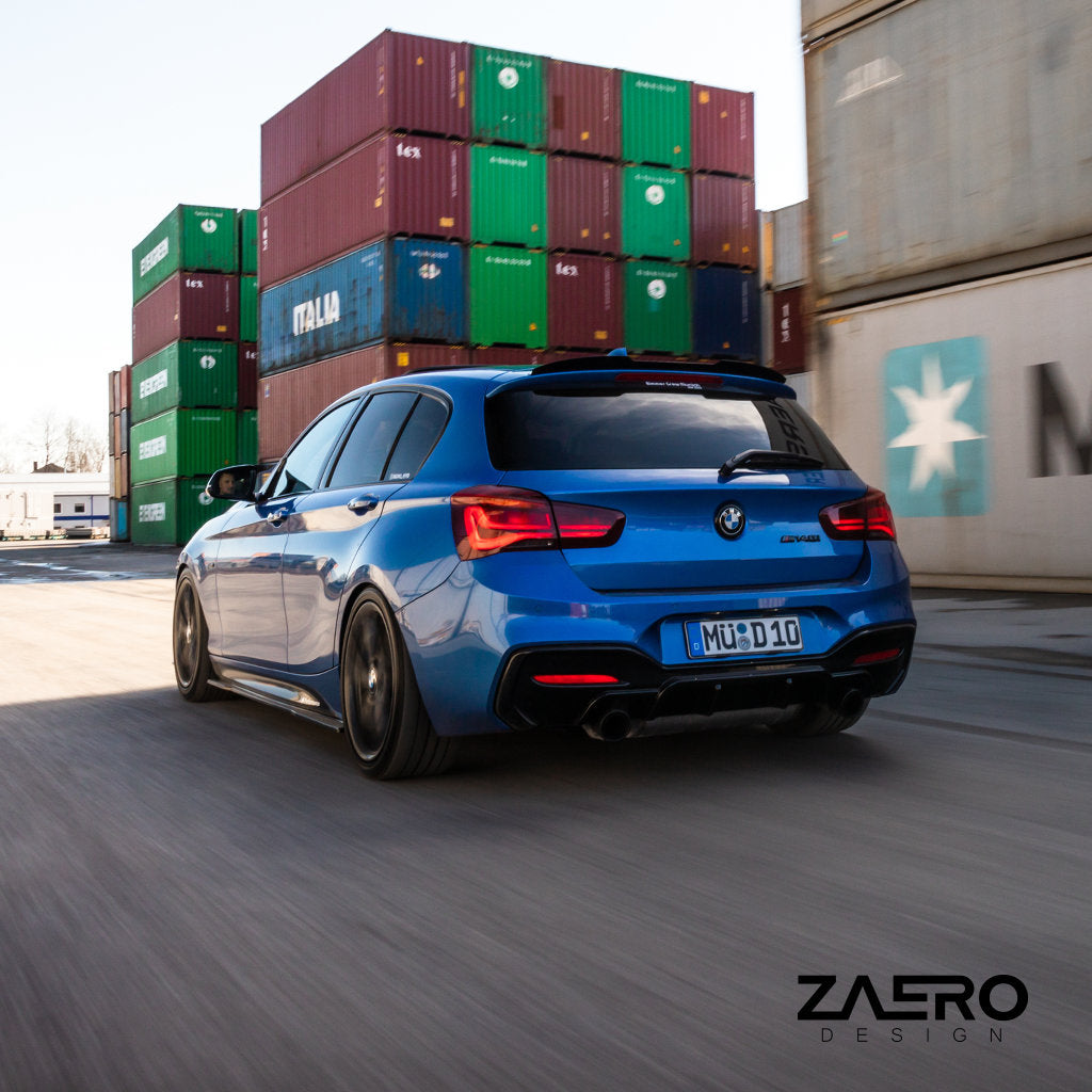 Ein blauer BMW M135i/M140i F2x mit ZAERO Evo-1 Diffusor und Carbon-Anbauteilen fährt in einem Industriegebiet an gestapelten Schiffscontainern vorbei, von hinten gesehen.