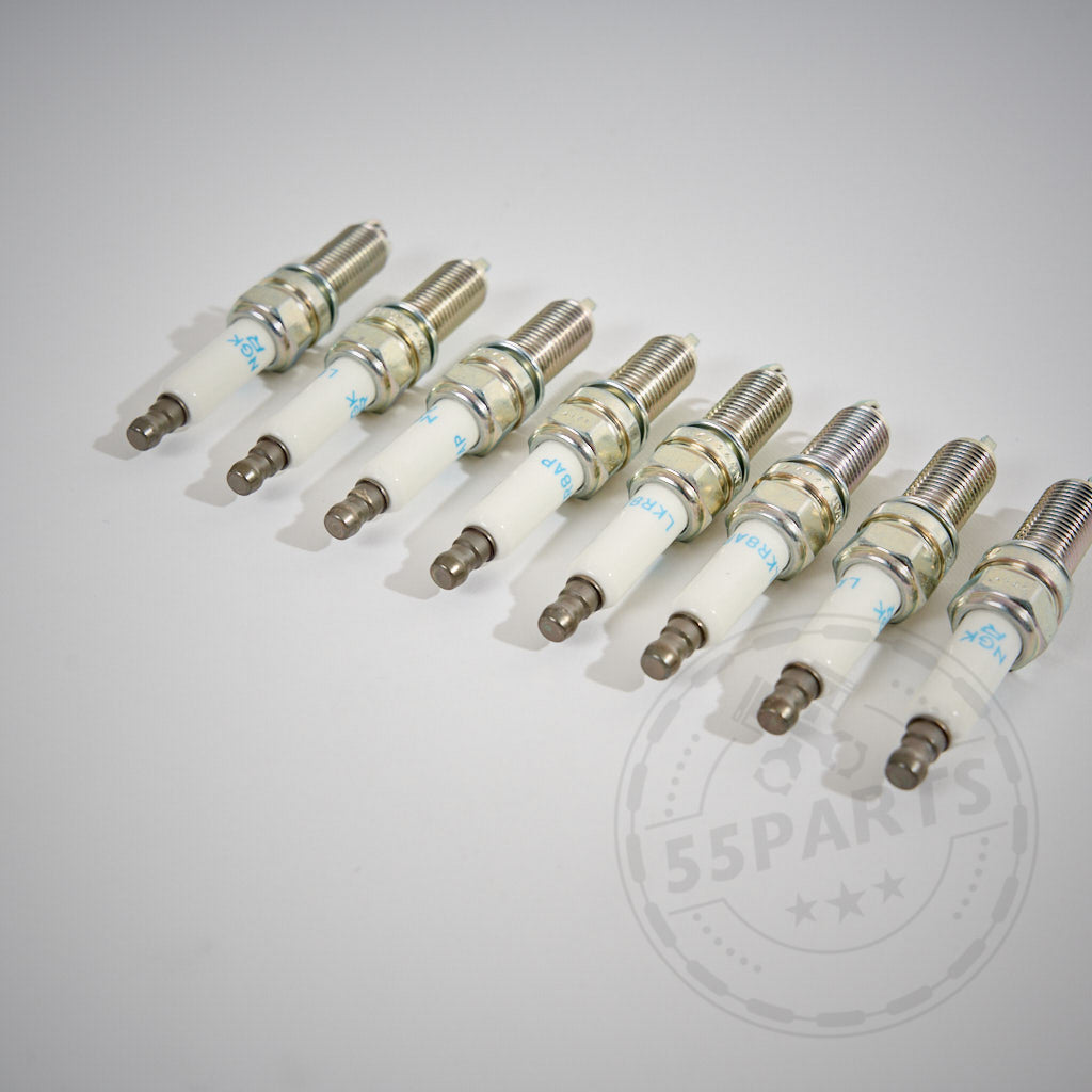 Ein Satz mit acht BMW Zündkerzen von NGK für den E9x M3 S65-Motor, diagonal angeordnet auf einer weißen Oberfläche mit einem schwachen PARTS-Logo-Wasserzeichen.