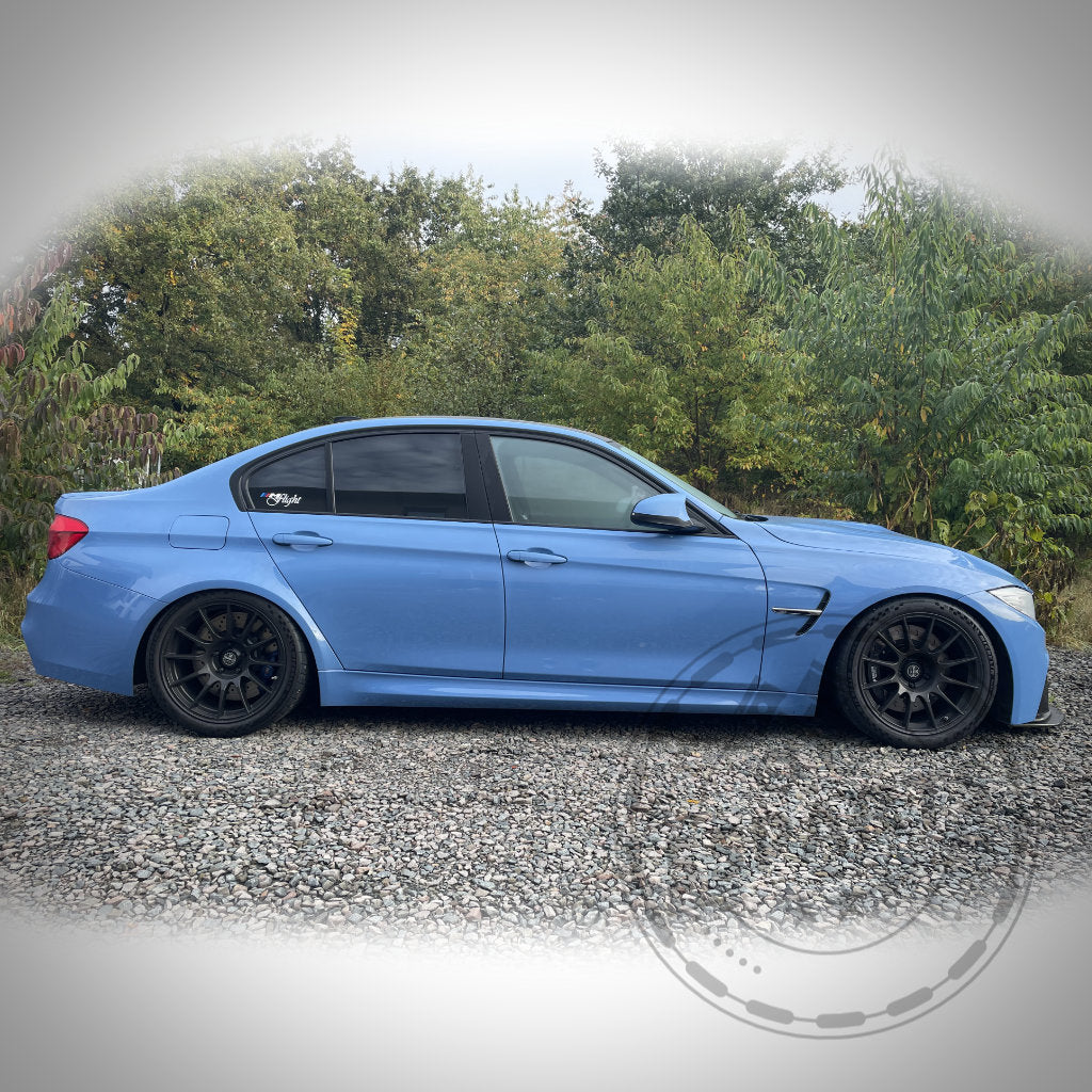 Eine blaue Sportlimousine mit CNC-gefrästen, verschraubten Radnabenkappen Special für ProTrack One-Räder ist auf Schotter geparkt, im Hintergrund sind Bäume zu sehen, mit einem weichen weißen Vignetteneffekt.