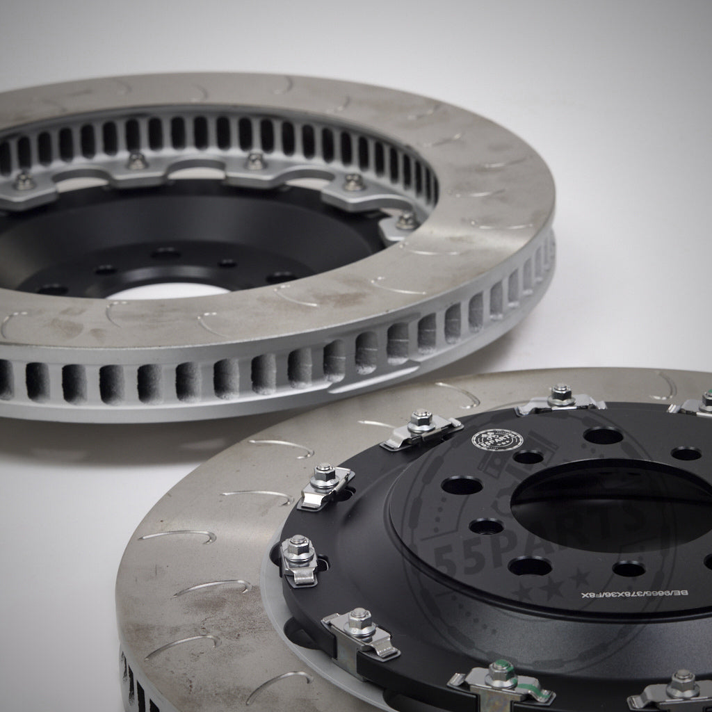 Auf weißem Hintergrund sind zwei belüftete, geschlitzte Bremsscheiben mit Metallbeschlägen abgebildet – perfekt zum Aufrüsten mit dem 55Parts-Special: AP Racing Big Brake Kit – Umrüstkit auf Aftermarket-Reibringe.