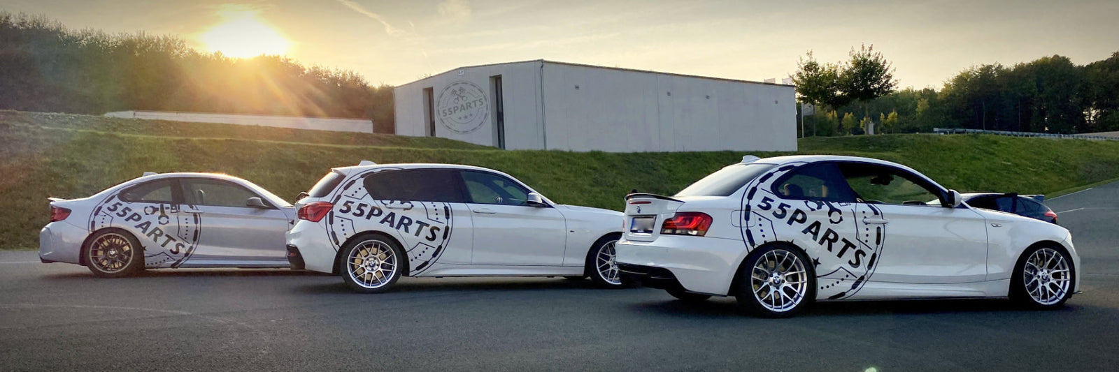Drei weiße Autos mit 5Parts-Aufklebern parken bei Sonnenuntergang auf dem Bürgersteig vor einem modernen Gebäude und einem grasbewachsenen Hügel.
