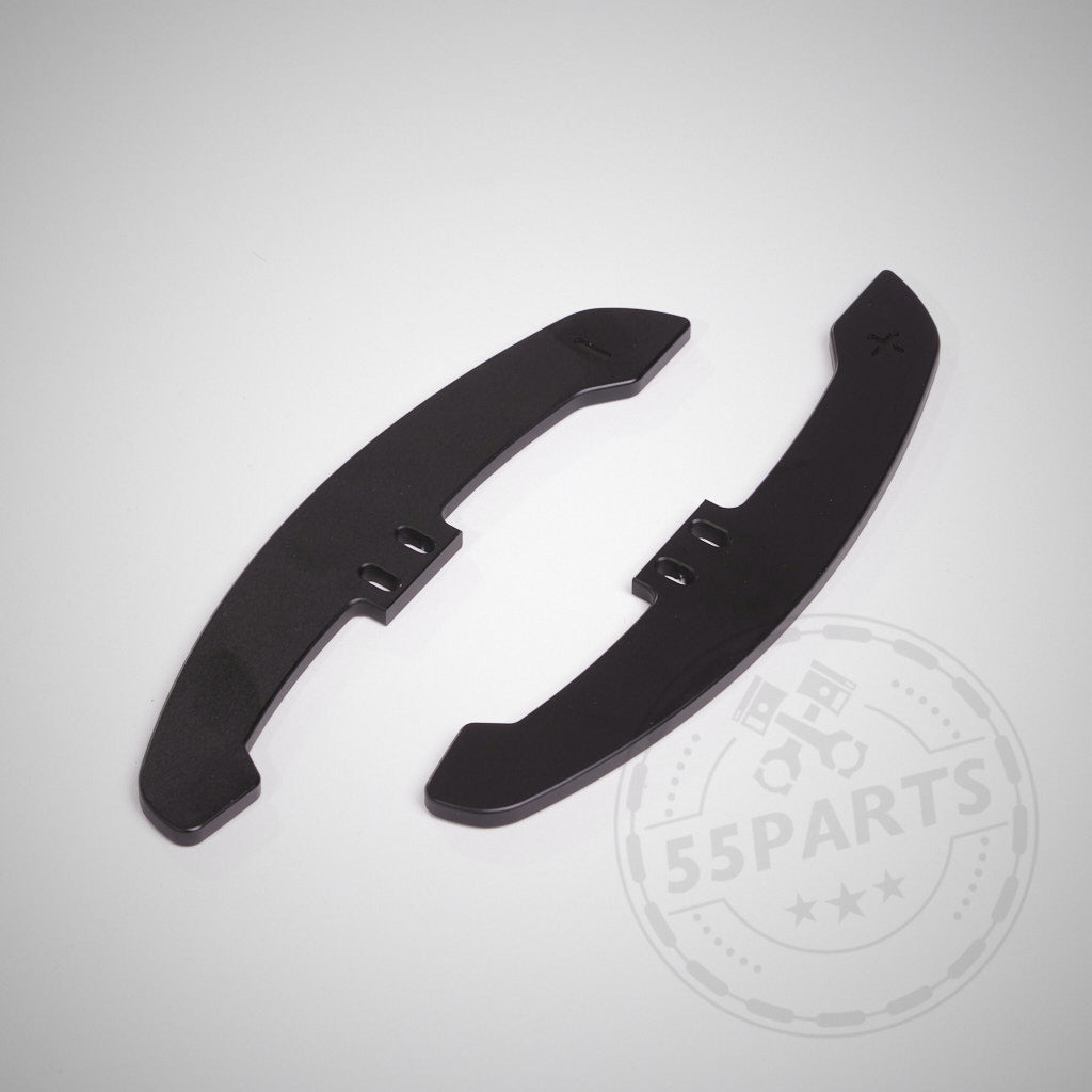 Zwei schwarze, gebogene 55Parts Special CNC-Aluminium-Schaltwippen für magnetische Schaltwippen sind auf einem hellgrauen Hintergrund mit einem schwachen 55Parts-Logo für magnetische Schaltwippen abgebildet.