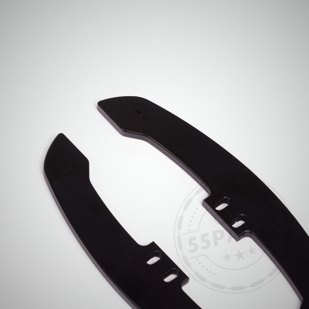 Zwei schwarze, CNC-gefräste magnetische Schaltwippen aus Aluminium Special mit Plus- und Minussymbolen liegen auf einem schlichten weißen Hintergrund.