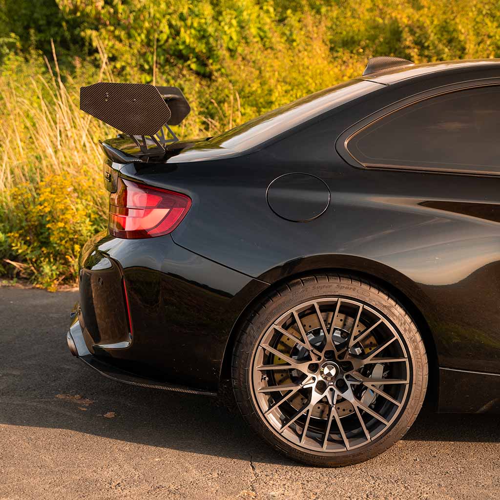 Schwarzer BMW M2 Competition Sportwagen mit einem 55Parts Motorsport Carbon Heckspoiler Heckflügel (passend für die meisten BMW Modelle, inklusive Teilegutachten), geparkt auf einer sonnenbeschienenen Straße, im Hintergrund Grünflächen.