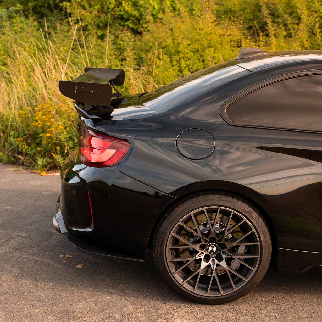 Heckansicht eines schwarzen BMW mit einem 55Parts Motorsport Carbon Heckspoiler Heckflügel, passend für fast alle BMW Modelle inklusive Teilegutachten, geparkt im hohen Gras an einem sonnigen Tag.