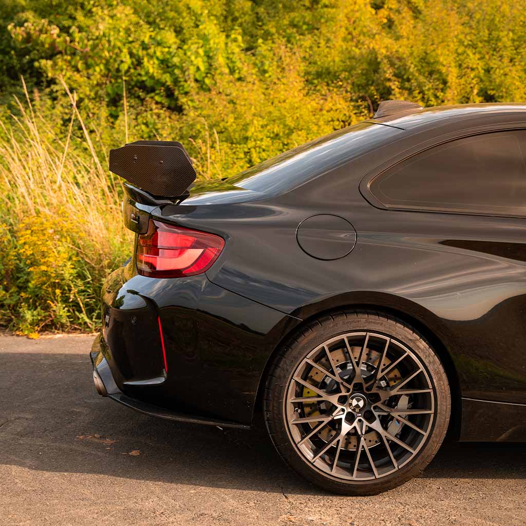 Nahaufnahme des Hecks eines BMW M2 Competition mit Rad, Rückleuchte und 55Parts Motorsport Carbon Heckspoiler Heckflügel (passend für die meisten BMW Modelle, inkl. Teilegutachten) vor üppiger grüner Kulisse.