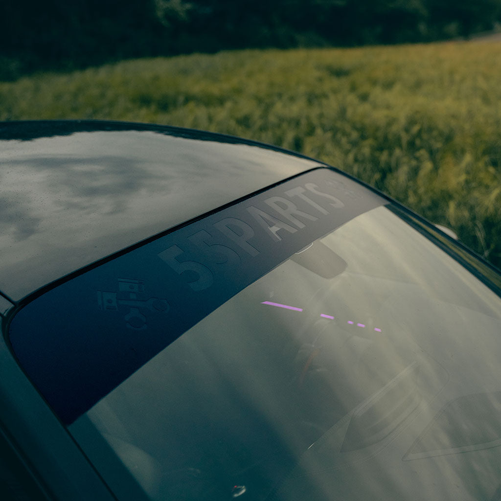 Ein 55Parts Scheibenkeil passend für alle Autos von 55Parts aus Oracal Markenfolie ist am Straßenrand neben einer Wiese unter bewölktem Himmel angebracht.