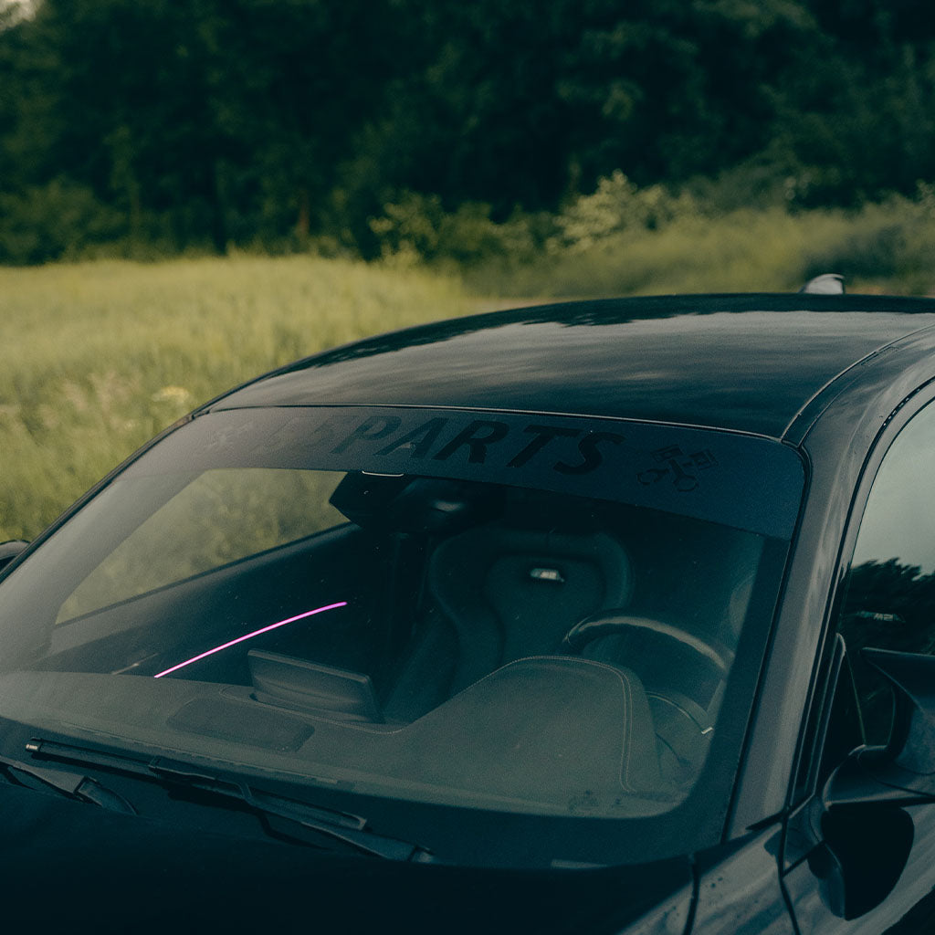 Durch die Windschutzscheibe ist ein schwarzes Auto zu sehen, das auf einer Wiese mit Bäumen geparkt ist. Der Scheibenkeil von 55Parts ist passend für alle Autos und sorgt für eine einfache, blasenfreie Montage.