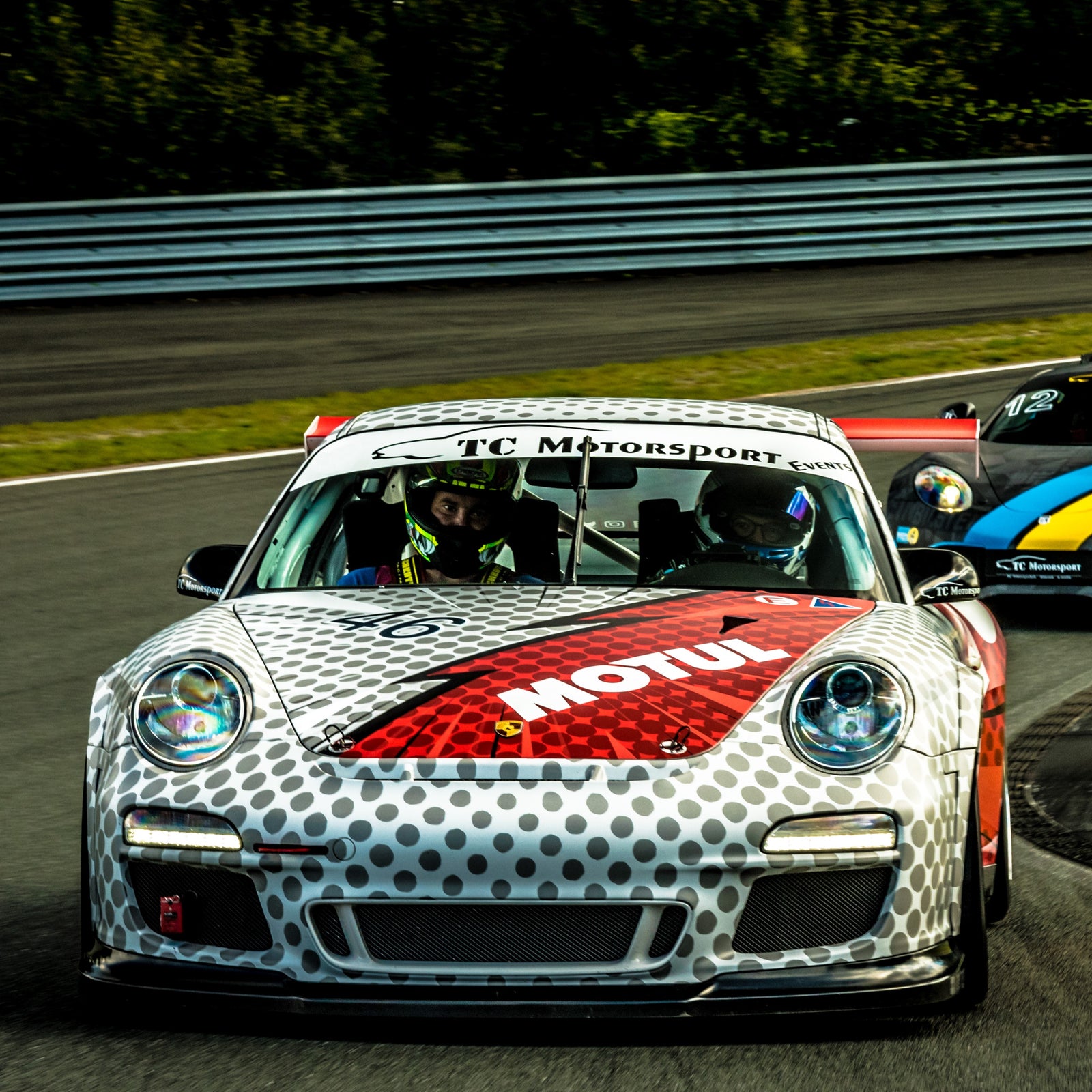 Ein Rennwagen mit Motul-Lackierung rast auf der Strecke davon, dicht gefolgt von einem anderen Fahrzeug. Beide werden von Rennfahrern mit Helmen gefahren.