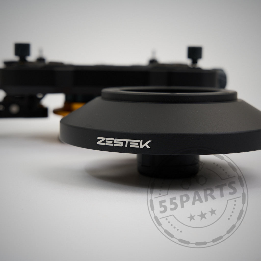 Im Fokus steht ein schwarzer Kameraobjektivadapter von Zestek, im Hintergrund sind verschwommene Geräte und das 55Parts Exklusiv: Zestek Lenkrad HUB für die BMW E/F/G-Serie und Toyota Yaris/Supra zu sehen.