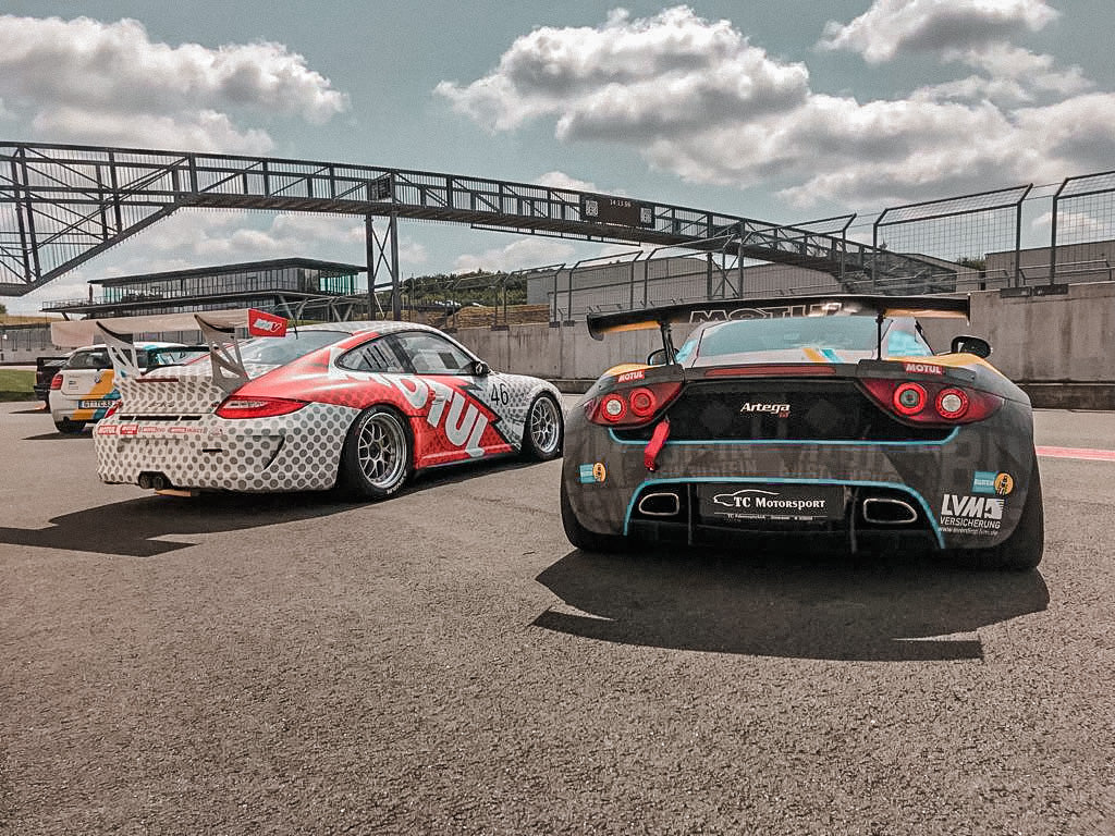 Zwei schnittige Rennwagen stehen unter einem teilweise bewölkten Himmel in der Nähe einer Boxengasse auf der Strecke und sind bereit für den Einsatz.