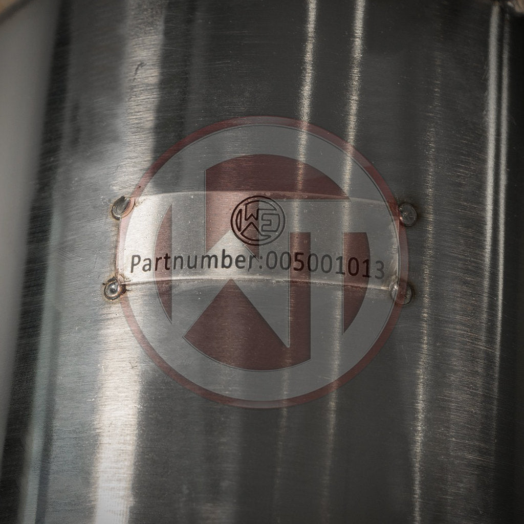 Eine Metallplatte auf glänzender Oberfläche mit der Teilenummer: 005001013, mit einem runden Logo im Hintergrund; geeignet für FBO-Setups oder als Wagner Tuning High Flow Downpipe mit 200 Zeller Kat passend für BMW 135i, 335i(x) N55 E-Serie.