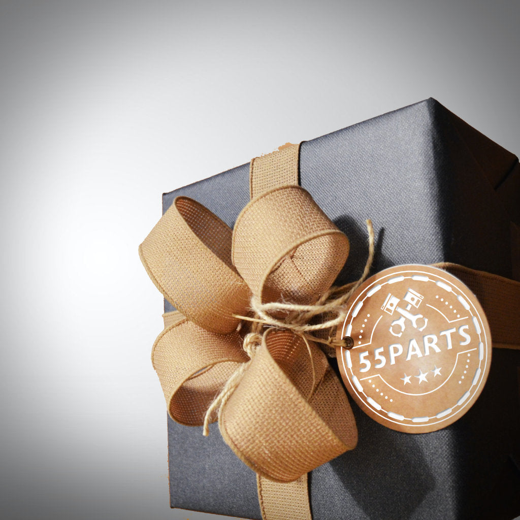Eine schwarze Geschenkbox mit beiger Schleife und rundem 55Parts-Anhänger liegt auf einem hellen Untergrund, ideal zur Präsentation des 55Parts-Geschenkgutscheins.