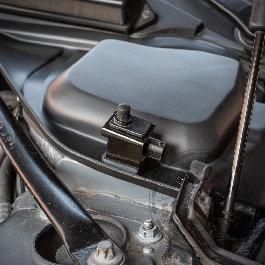 Nahaufnahme des Bannik Engineering Mikrofiltergehäuse-Unterteils (für bessere Wärmeabfuhr) im BMW Motorraum, passend für E8x/E9x 1er und 3er, mit sichtbaren Metallteilen im Hintergrund.