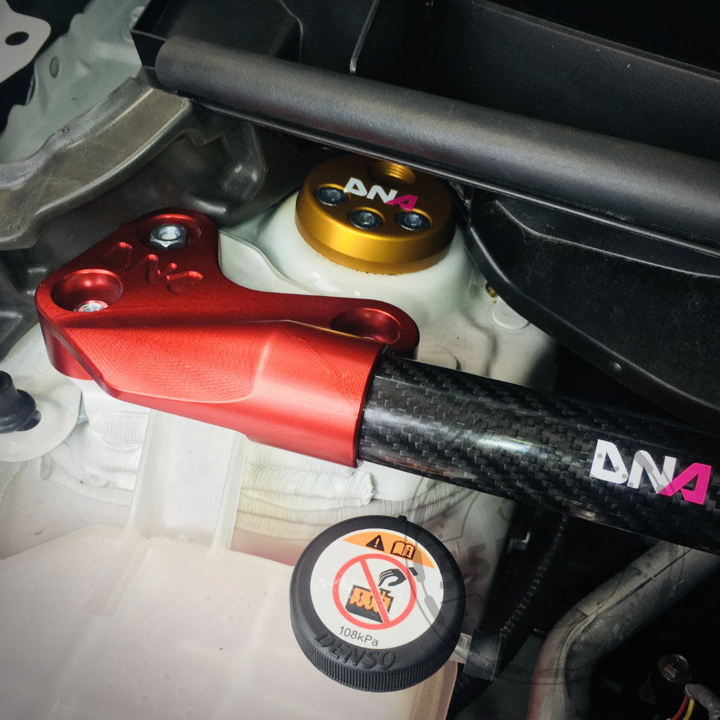 Nahaufnahme eines Yaris GR-Motorraums mit verstellbarem Uniball-Domlager von DNA Racing, einer rot-schwarzen Domstrebe von DNA Racing, Flüssigkeitsbehältern und sichtbaren Fahrwerk-Tuning-Komponenten.