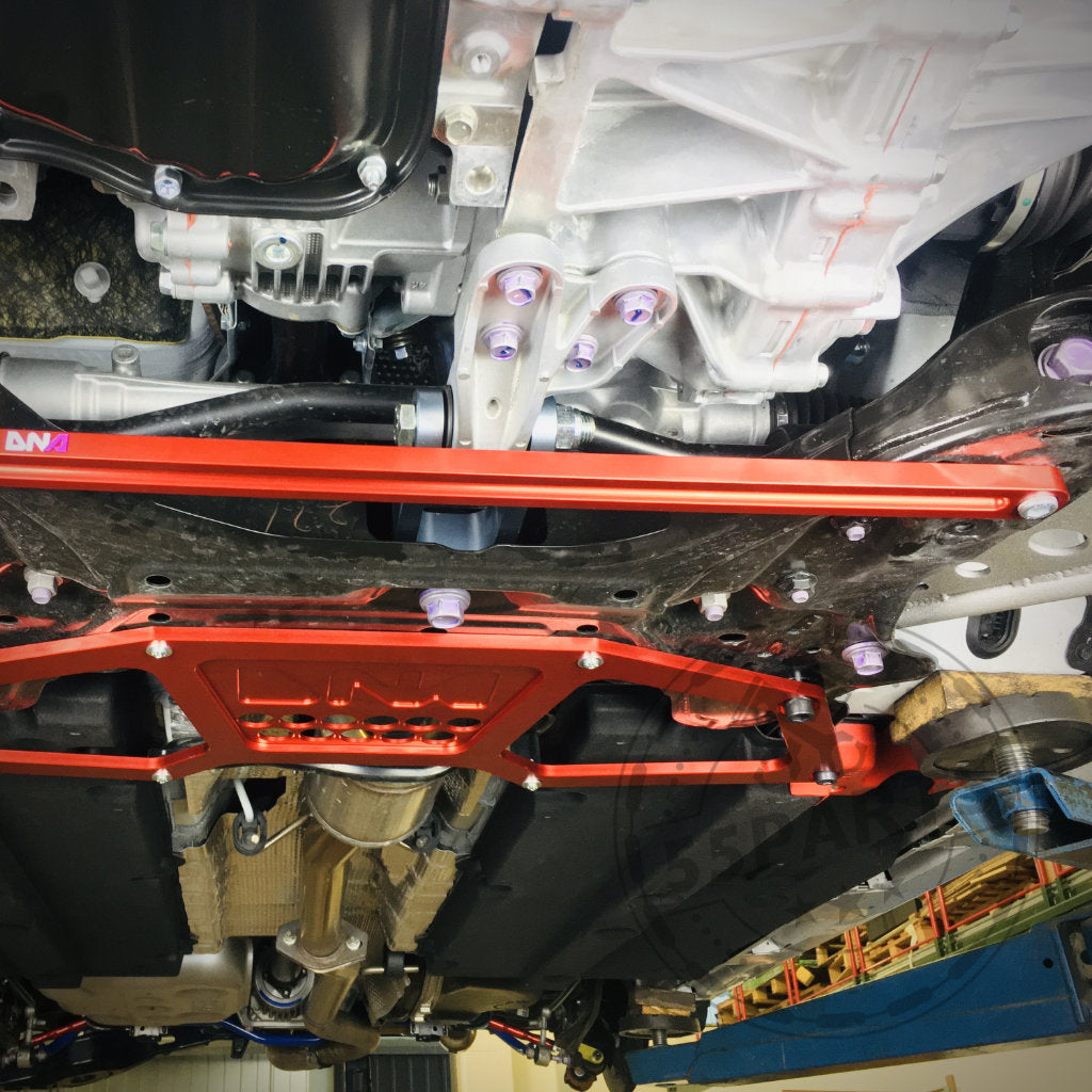 Unterseite eines Yaris GR auf einer Hebebühne, die die Versteifungsstrebe vorderer Achsträger vorne von DNA Racing zeigt, eine vordere Aufhängungsstrebe von DNA Racing, die zwischen Metallkomponenten montiert ist.