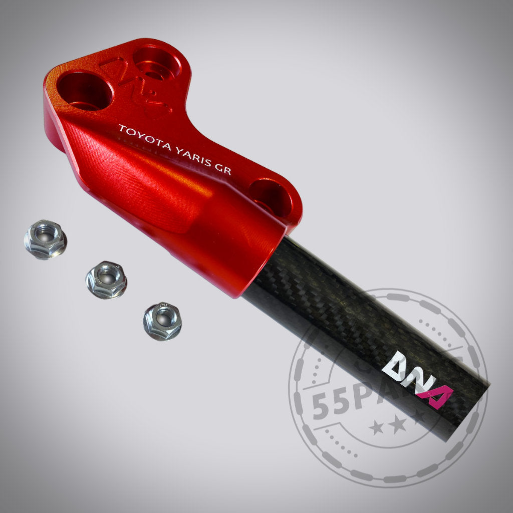Die DNA Racing Carbon Domstrebe vorne für den Toyota Yaris GR verfügt über eine schwarze Carbonfaserstrebe mit roten Metallhalterungen, ideal für Fahrwerks-Upgrades, und ist auf einem hellgrauen Hintergrund abgebildet.