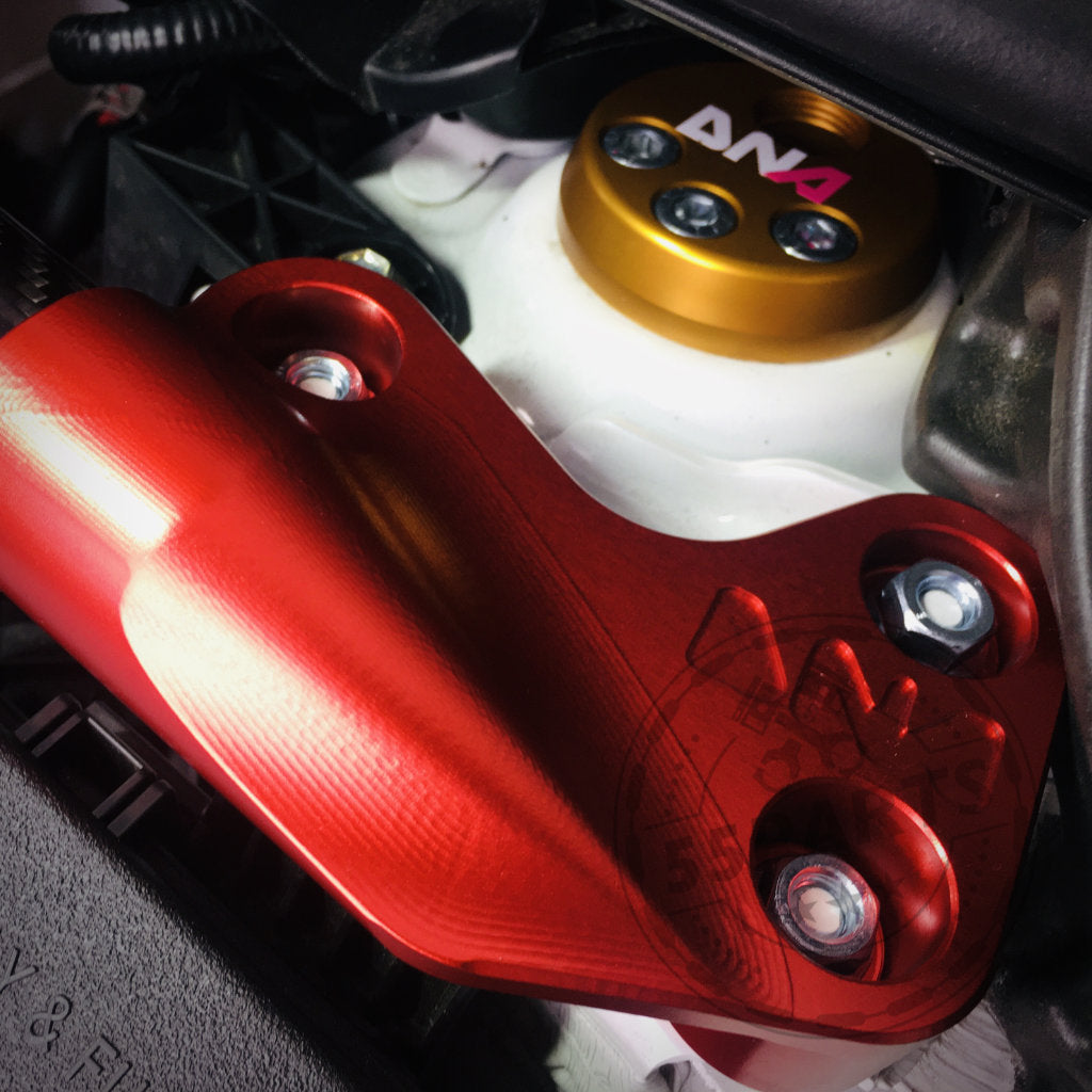 Nahaufnahme der DNA Racing Carbon Domstrebe vorne für den Toyota Yaris GR. Sie zeigt ein rotes Metallteil mit Schrauben und eine goldene AN4-Kappe auf einer weißen Motorkomponente, ausgestellt in einem Tuning-Shop.