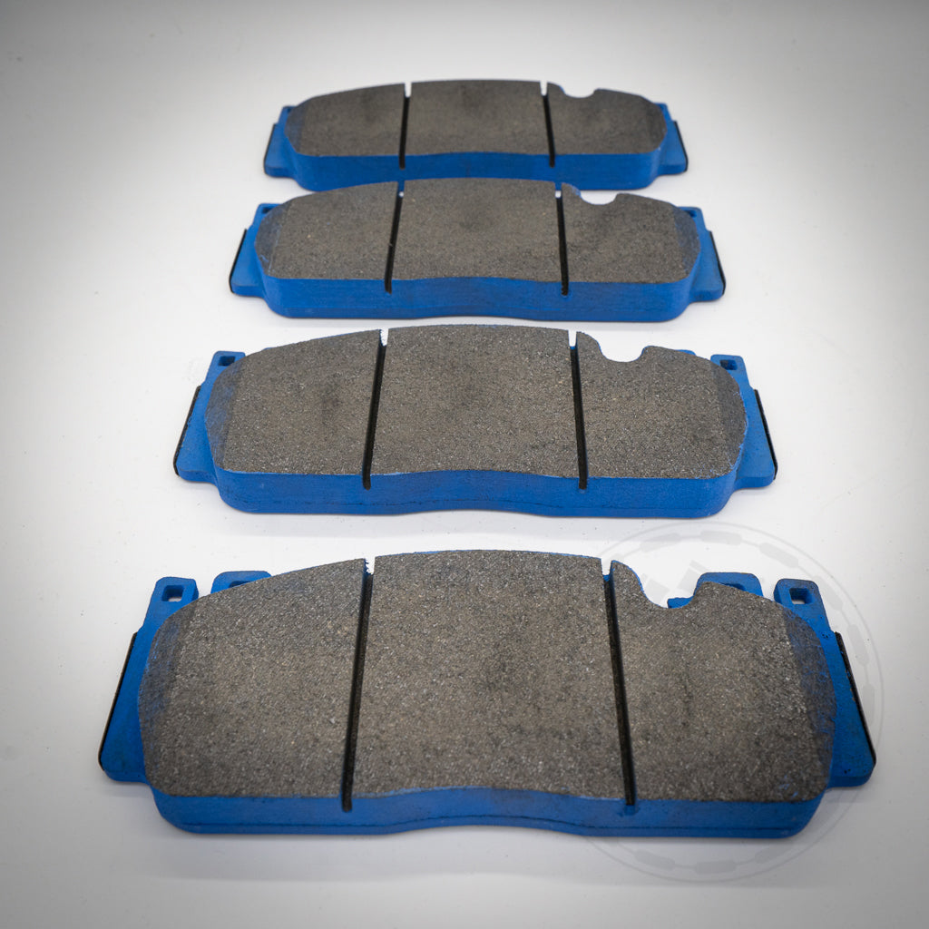 Vier Endlos-Bremsbeläge mit blauer Rückseite passend für AP Racing Pro5000R Bremssattel sind in einer Reihe auf einer weißen Fläche angeordnet, aus einem niedrigen Winkel betrachtet.