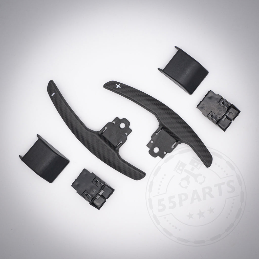Auf hellgrauem Untergrund sind zwei 55Parts Aluminium/Carbon Schaltwippen für die BMW F- und G-Serie und vier schwarze Kunststoffteile angeordnet.