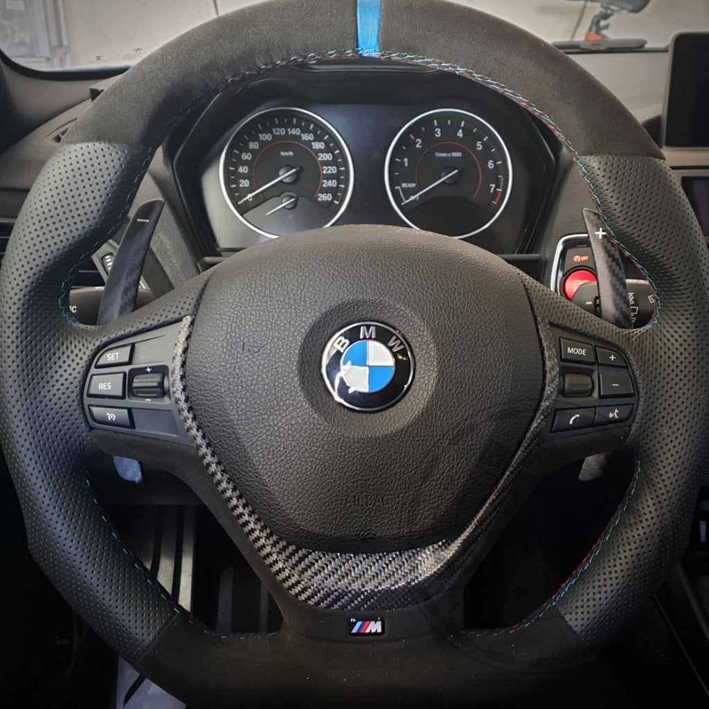 Nahaufnahme der 55Parts Aluminium/Carbon-Schaltwippen passend für BMW 1/235/40i, 3/435/40i, M2, M3 und M4 F- und G-Serie auf einem M-Lenkrad mit Bedientasten und Armaturenbrettanzeigen im Hintergrund.