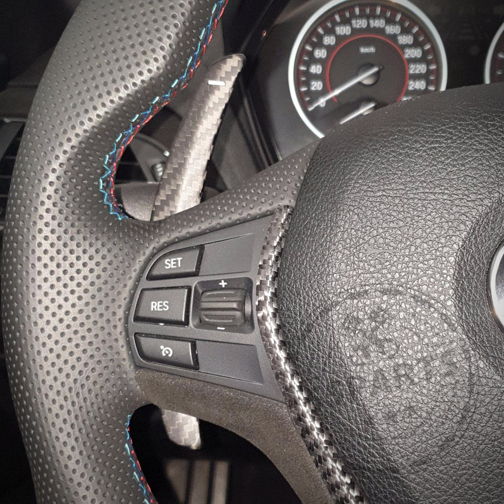 Nahaufnahme der Schaltwippen und Steuertasten Aluminium/Carbon an einem Lenkrad eines BMW 1/235, 40i, 3/435, M2, M3 oder M4 (F-/G-Serie), mit Tachometeranzeigen im Hintergrund.