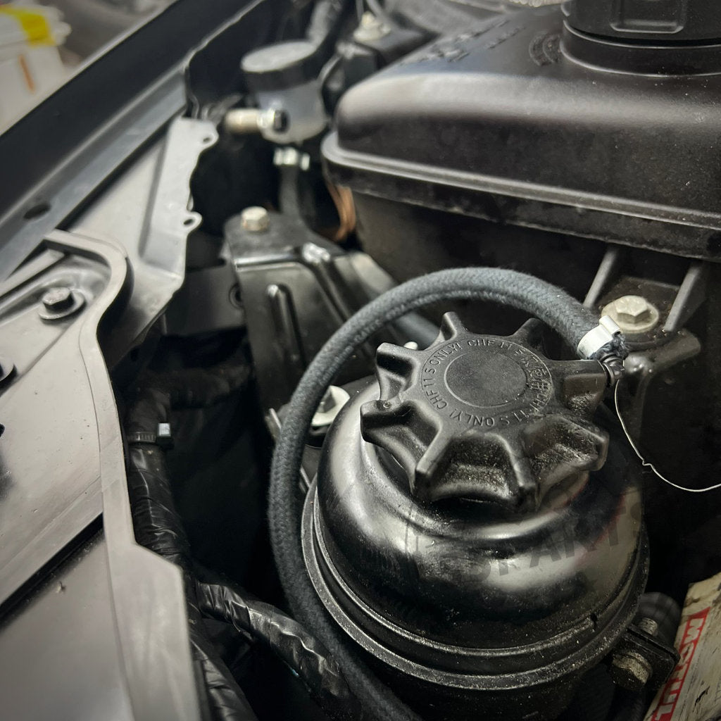 Nahaufnahme eines 55Parts Spezial-Ausgleichsbehälters/Catch Tank für Hydrauliköl Servoflüssigkeit, passend für BMW 3er E9x und 1er E8x inkl. M3, mit Kappe und Schlauch zwischen Motorteilen aus Metall und Kunststoff.