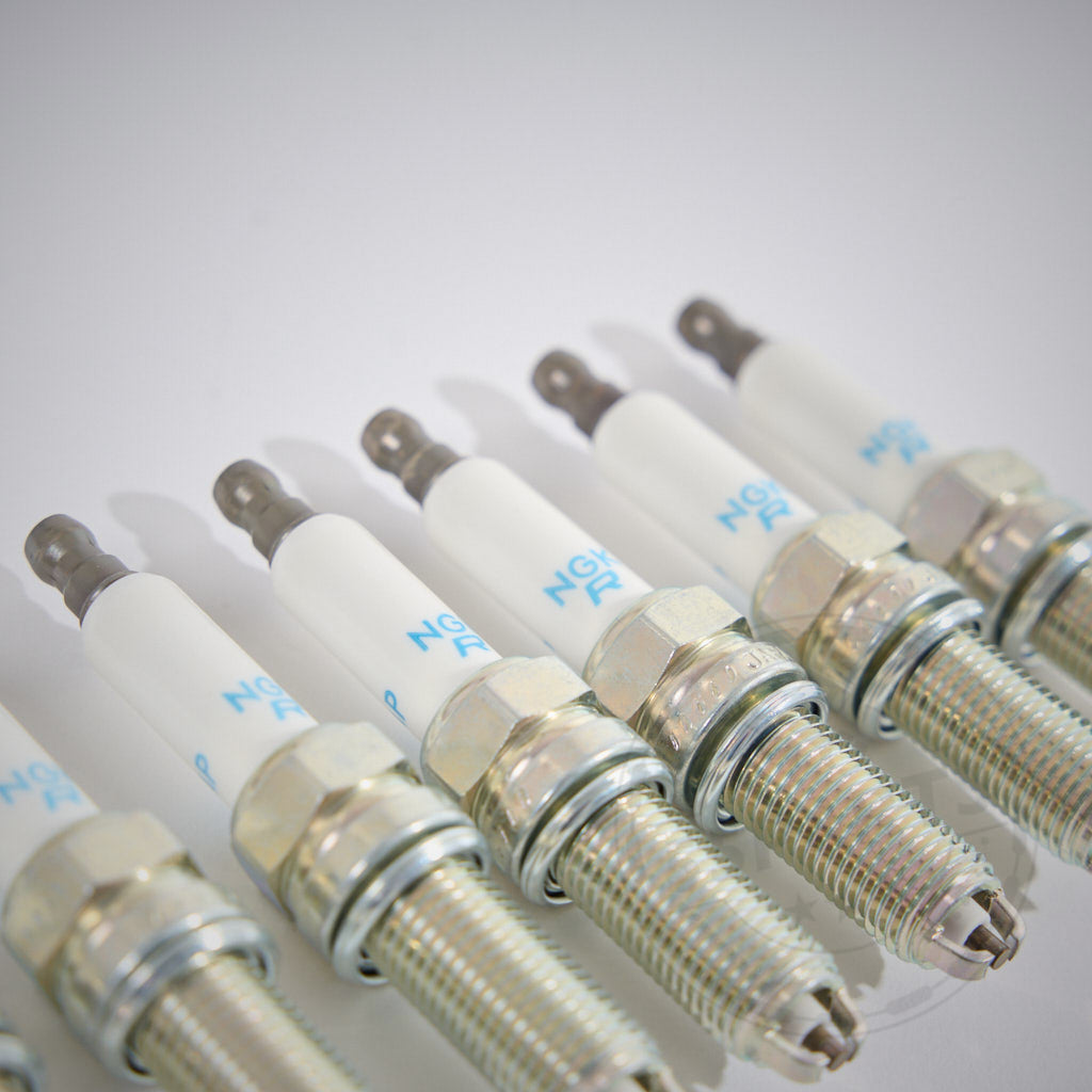 Sechs Zündkerzen aus dem BMW Zündkerzen Set NGK für 8-Zylinder E9x M3 S65 Motoren sind diagonal auf einer weißen Fläche angeordnet und heben ihr metallisches und keramisches Design hervor.