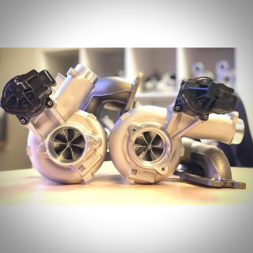 Zwei silberne Pure Stage 2 Upgrade Turbolader von Pure Turbos passend für BMW M2 Competition, M3 F80 und M4 F8x S55 mit angebauten schwarzen Sensoren werden auf einer hellen Fläche mit unscharfem Hintergrund dargestellt.